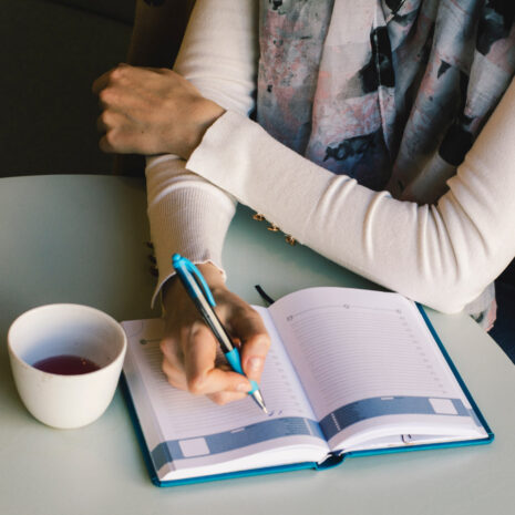 A woman sits a table with a hot drink, journalling in her book and looking very relaxed.
