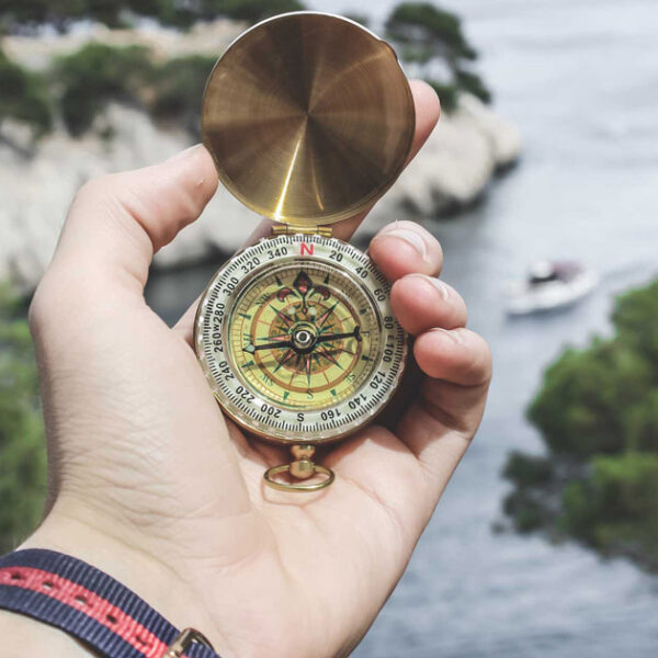 A person holds up a compass in front of them by the coastlline to see which way is North.
